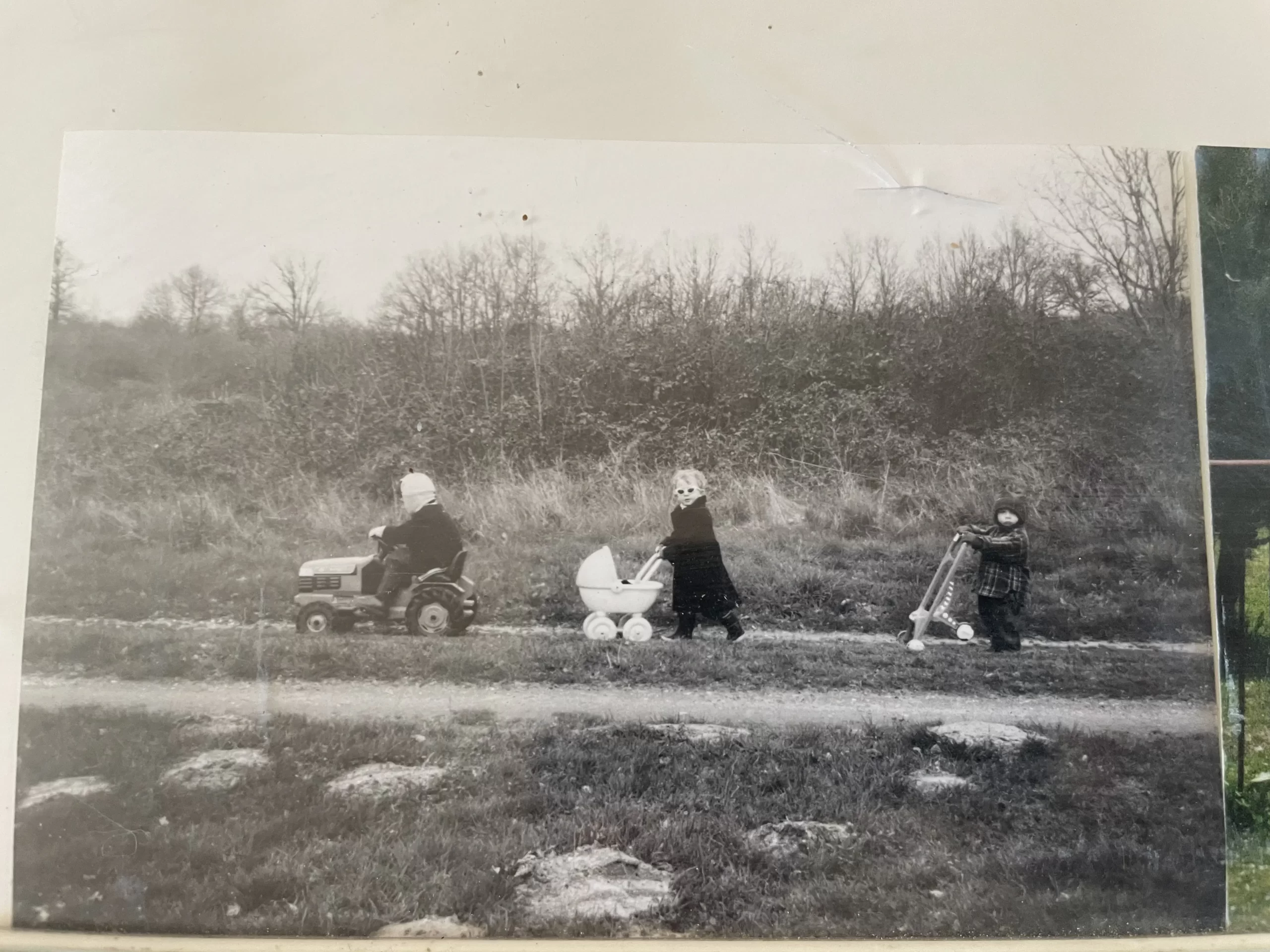 Three children walking along a countrypath. Frist child on a tractor in a hat very concentrated. Second child, female pushing a pram and is looking at the camera - has sunglasses on. Third child, male, is lifting the pushchair and stopped.